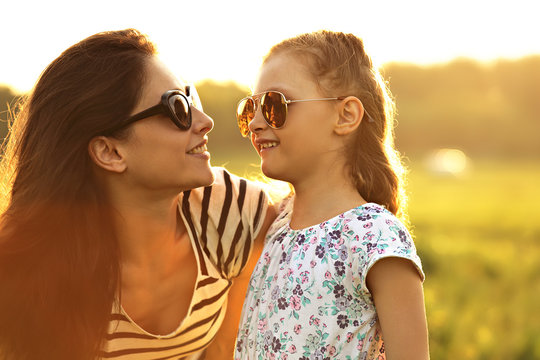 Happy fashion kid girl and her mother in trendy sunglasses looking on each other on nature sunset background. Closeup portrait of happiness.