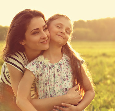 Emotional calm mother hugging her enjoying kid girl with closed eyes on sunset bright summer background. Closeup toned soft color portrait