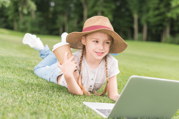 happy child holding paper cup and using laptop while lying on grass in park