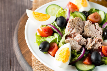 Fresh fish tuna salad made of tomato, ruccola, tuna, eggs, arugula, crackers and spices. Caesar salad in a white bowl on wooden background