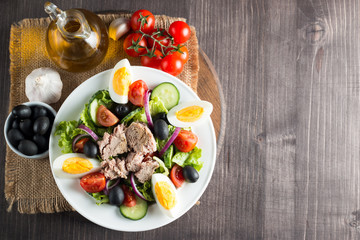 Fresh fish tuna salad made of tomato, ruccola, tuna, eggs, arugula, crackers and spices. Caesar salad in a white bowl on wooden background