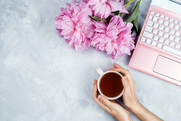 Feminine business mockup with pink laptop, peonies bouquet and woman's hands holding cup of warm...