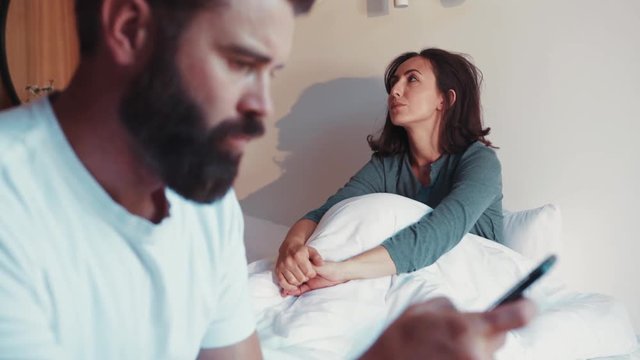 Disappointed bearded man sits on the corner of the bed, checks his wife&rsquo;s phone, his irritated wife sits on the background. Jealousy, conflict situation, no trust. Fighting, arguing. Bad mood.