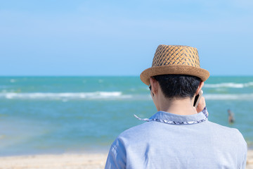  Rear view and copy space of Asian man wearing a hat. Talking on the phone is on the beach outdoors during the daytime.