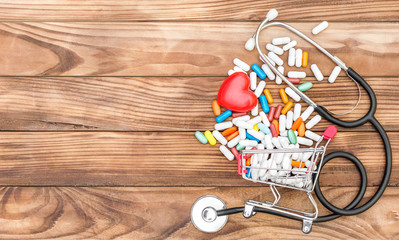 Shopping cart with pills with red heart and stethoscope on wooden background. Space for text. Medical background. Top view.