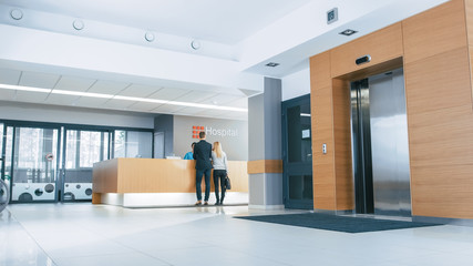 In the Hospital Lobby Young Couple Patients Talks to Receptionist. New, Modern Medical Facility.