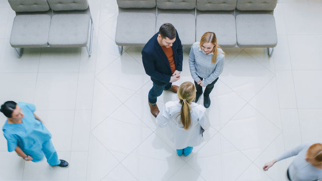 High Angle Shot In The Hospital Lobby, Young Couple Talks Meets With  Female Physician / Doctor / Nurse, They Have Discussion. New, Clean, Modern Hospital With Friendly And Professional Personnel.