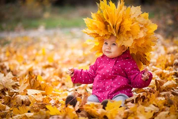 Poster Klein kind speelt en zit in gevallen bladeren in het herfstpark. Baby is in grote krans van bladeren. Meisje is gekleed in warme muts, jas. © Marina April