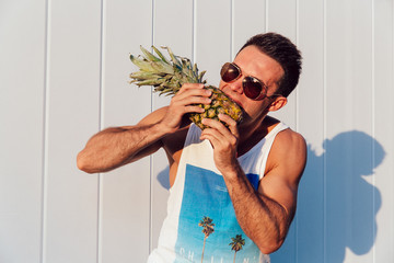 Stylish muscular man wearing sunglasses trying to eat pineapple, standing outdoors. Summer time...