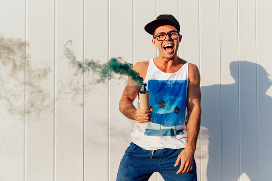 Stylish smiling man in eyeglasses holding green smoke bomb, standing near the urban wall, outdoors. Dressed in fashionable singlet, shorts and cap.