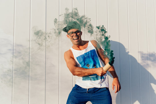 Trendy funny man having fun outside, holding green smoke bomb, enjoying the summer days, wearing fashionable clothes, eyeglasses and cap.