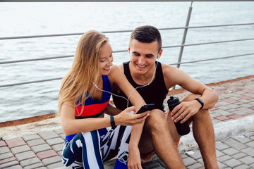 Happy sportswoman and smiling sportsman looking at smartphone screen and watching a funny video in headphones, while sitting on the ground and resting after workout. On the quay, near the ocean.