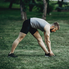 Muscular man runner doing stretching exercise, preparing for morning workout in the park. Street fitness, sport, exercising, people and healthy lifestyle concept.