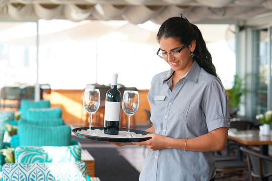 A young waitress carries a bottle of red wine with wine glasses on a tray to the customer of the hotel restaurant. The concept of service.