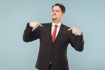 A very proud man shows his fingers and boasts. indoor studio shot. isolated on light blue background. handsome businessman with black suit, red tie and mustache looking away.