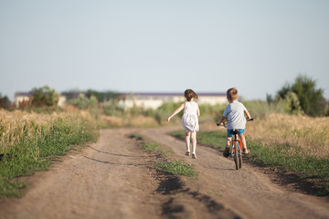  Children are walking in the field. The boy is riding a bicycle. The girl runs next to him.