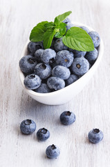 Drops fresh blueberries in a white bowl, with meant leaves on a wooden background