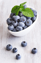 Drops fresh blueberries in a white bowl, with meant leaves on a wooden background