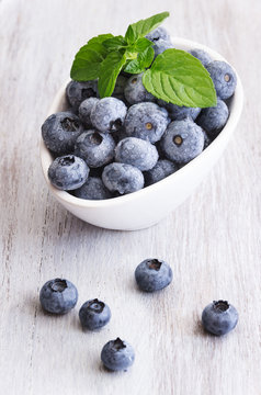 Drops fresh blueberries in a white bowl, with meant leaves on a wooden background