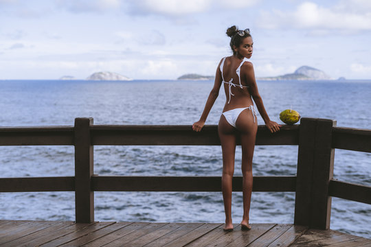 Rear view of a charming young black girl standing half-turned to the camera in the swimsuit on the observation point near the ocean with coconut next to her; copy space area on the left for a text