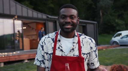Close up attractive african american man cook food smile look at camera happy in background barbecue for summertime dinner party girl couple play guitar rest outdoors sunset slow motion portrait