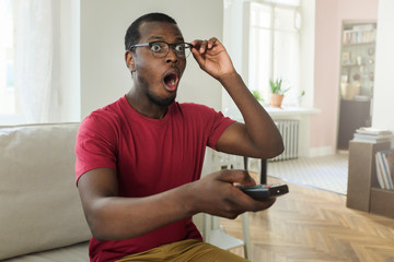 Portrait of young African American man sitting on couch in his living room wearing casual T-shirt,...
