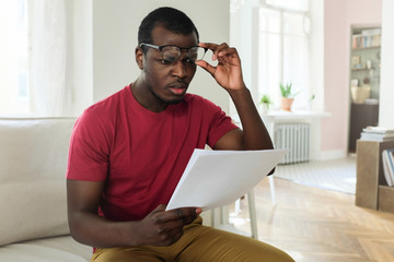 Horizontal picture of handsome African American man sitting on sofa with sheets of paper in hand looking at numbers in bills from under glasses feeling dissatisfied with his financial problems