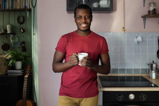 Horizontal closeup of young African American man standing in casual clothes in kitchen holding white cup of hot drink smiling confidently to person he is talking to, enjoying his leisure time