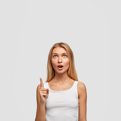 Fototapeta na wymiar Vertical shot of surprised young female opens mouth and looks upwards with astonished expression, wears white casual vest, points up, has attractive appearance, isolated over white background