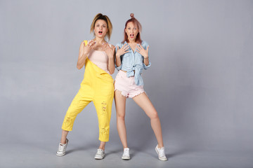Fototapeta na wymiar Full length of two cheerful young female in color casual demin clothes having shocked and scared expressions, mouths wide open, feeling nervous and frightened. People emotions