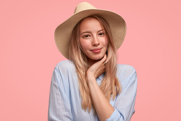 Portrait of attractive lovely young female wears elegant hat and shirt, holds hand under chin, shows her natural beauty, stands against pink background. People, feminity and fashion concept.