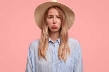 Discontent lovely female tourist in elegant hat, purses lower lip, feels lonely or abused, doesn`t have companion to travel, poses against pink background. Lonely sorrowful young blonde woman