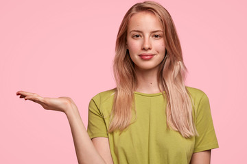 Cute young female with pleasant appearance, raises palm, advertises something, dressed in green...