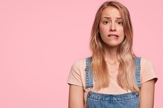 Embarrassed puzzled lovely female bites lips, has miserable facial expression, dressed in fashionable denim overalls, stands against pink blank wall. People, reaction, feeling and doubt concept