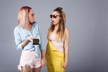 Young girls in fashionable colored casual clothes fool around looking at each other, using smartphone and social networks. Portrait of female best friends makes funny grimaces, show tongue, smiling