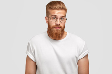 Puzzled ginger male freelancer feels surprised, looks in bewilderment, wears round spectacles,...