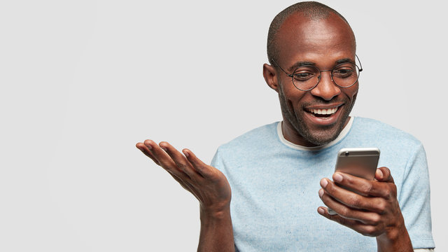 Horizontal portrait of handsome cheerful African American male looks happily into smart phone, raises palm, wears glasses, isolated over white background with copy space for your text. Technology