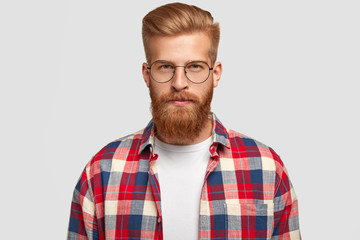 Fototapeta premium Studio shot of ginger male hipster in eyewear and checkered shirt, looks serious with confident facial expression, recieves necessary information, isolated over white background. People and style