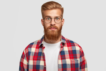 Obraz premium Portrait of handsome young successful male designer with thick ginger beard, looks surprisingly at camera, has serious expression, wears big round spectacles and checkered shirt, stands indoor