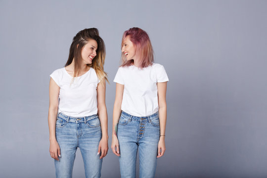 Horizontal portrait of two pretty women with happy expression, wears similar casual white t-shirt with denim, poses over gray background. Adorable lovely female student smile at each other. Copyspace