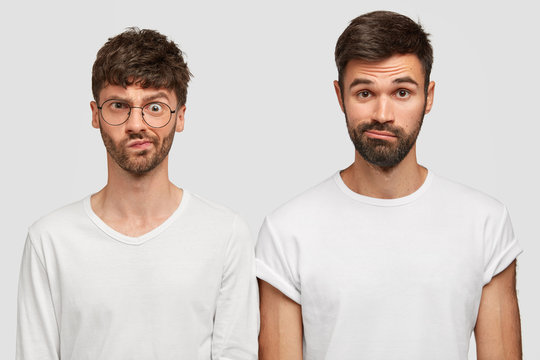 Horiozntal shot of displeased two men frown faces in discontent, have puzzled expressions, dressed casually, have thick stubbles. Male students wait for results of exam, isolated on white wall
