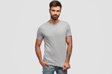 Fototapeta premium Stylish bearded handsome male with thick dark stubble, dressed in casual t shirt, keeps hand in pocket of jeans, looks self assured, fond of fashion, isolated over white background. Masculinity