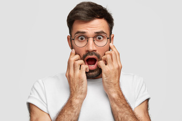 Horizontal shot of attractive bearded male stares with frightened expression, keeps hands near mouth, can`t beleive his eyes, notices something awful on road, stands alone against white concrete wall