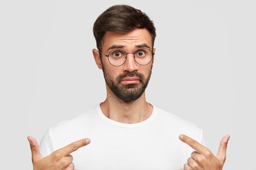 Emotive young handsome male with thick dark beard, points at blank t shirt with bewilderment, shows blank space for your logo or advertisement, stands against white background. Clothing concept