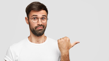 Indoor shot of displeased bearded male with doubtful surprised expression, points aside with thumb, shows free space for your promotional text or advertisement. People and facial expressions