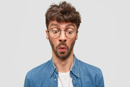 Indoor shot of surprised unshaven male wonders while notices something down, has displeased expression, wears glasses and shirt, stands alone against white background. Omg and reaction concept