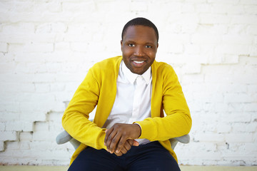 Obraz premium People and lifestyle concept. Studio shot of happy positive young Afro American guy sitting on chair and looking at camera with broad joyful smile, showing his white perfect teeth, clasping hands