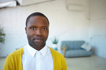 Close up shot of attractive young unshaven African American male wearing trendy yellow cardigan over white shirt, looking and smiling at camera, standing in modern living room with sofa in background