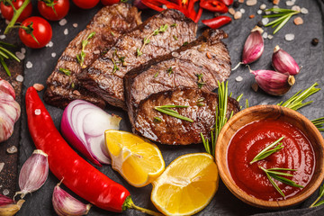 Grilled beef steaks with spices and vegetables on slate board, dark background