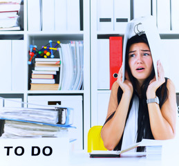 Funny overworked girl with a folder on her head sitting in the workplace cluttered with folders....